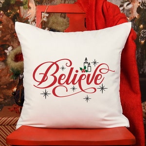 May include: White pillow with the word "Believe" in red cursive lettering, with a small image of candles and holly berries above the word. The pillow is on a red chair in front of a Christmas tree.
