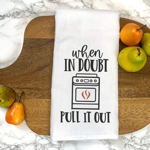 May include: White kitchen towel with black text that reads "When in doubt, pull it out."  A black and white illustration of a stove with a red flame is above the text. The towel is on a wooden chopping board with pears.