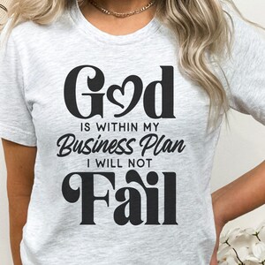 May include: A white t-shirt with black text that reads "God is within my Business Plan I will not Fail".