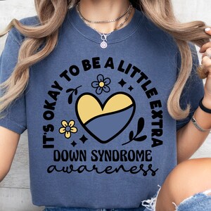 May include: A blue t-shirt with the text "It's okay to be a little extra Down Syndrome awareness" in a circle with a yellow and blue heart and flowers.