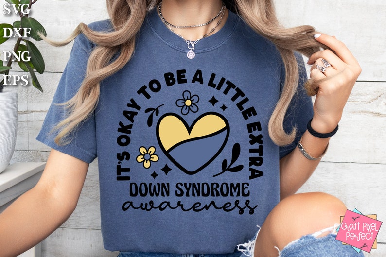 May include: A blue t-shirt with the text "It's okay to be a little extra Down Syndrome awareness" in a circle with a yellow and blue heart and flowers.