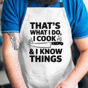 May include: A white apron with black text that reads "That's what I do, I cook & I know things". A knife with flames on either side is pictured above the text. A person wearing the apron is standing in front of a white wall.