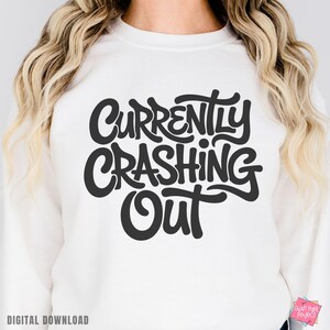 May include: White sweatshirt with the phrase "Currently Crashing Out" in large, black, stylized lettering. The words are in a bold, rounded font. The phrase "Digital Download" is at the bottom left.