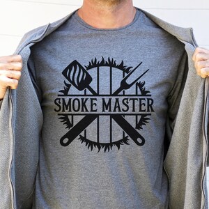 May include: A gray t-shirt with a black graphic design of a grill with crossed grilling tools and flames. The text "SMOKE MASTER" is in the center of the design.