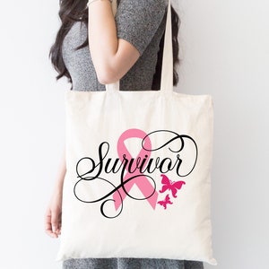 May include: A white tote bag with a pink ribbon and the word "Survivor" in black cursive script. Two pink butterflies are on the right side of the bag.