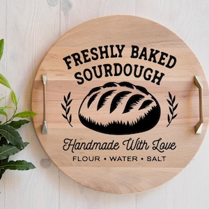 May include: A circular wooden serving board with handles, displaying the text "FRESHLY BAKED SOURDOUGH" and a bread illustration. The text "Handmade With Love" and ingredients are also included. The board is light brown.