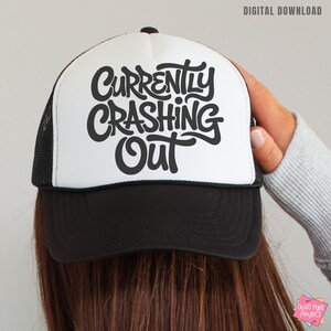 May include: A black and white trucker hat with the words "Currently Crashing Out" in a bold, stylized font. The hat has a white front panel, a black brim, and a black mesh back. The text is in black.