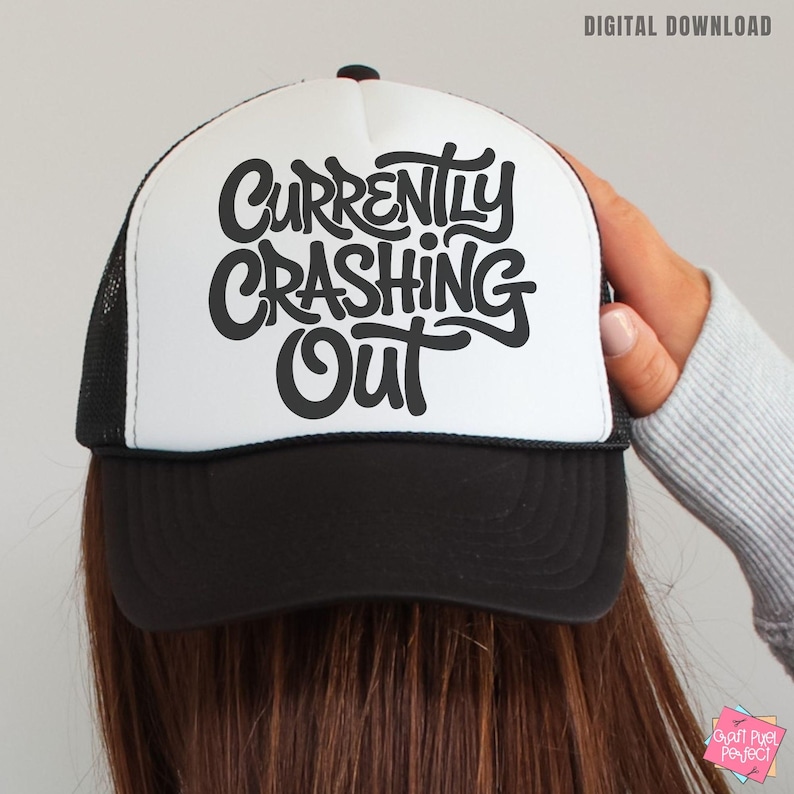 May include: A black and white trucker hat with the words "Currently Crashing Out" in a bold, stylized font. The hat has a white front panel, a black brim, and a black mesh back. The text is in black.