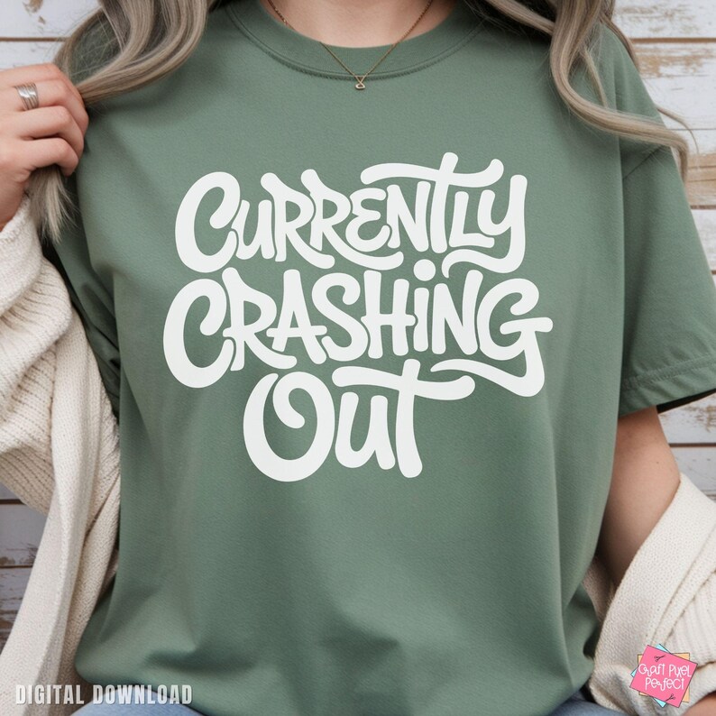 May include: Sage green t-shirt with the white text "Currently Crashing Out" in a playful, rounded font. The shirt is a relaxed fit, with short sleeves. The image shows a close-up of the shirt.
