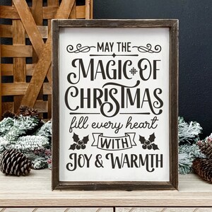 Puede incluir: Un letrero de madera con fondo blanco y texto negro que dice "Que la magia de la Navidad llene cada coraz&oacute;n de alegr&iacute;a y calidez". El letrero est&aacute; decorado con acebo y una cinta.
