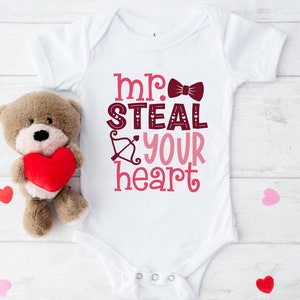 May include: White baby onesie with the text "mr. steal your heart" in pink and burgundy lettering with a bow tie graphic. A teddy bear holding a red heart is to the left of the onesie.