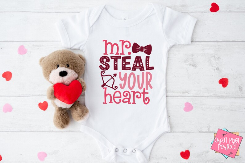 May include: White baby onesie with the text "mr. steal your heart" in pink and burgundy lettering with a bow tie graphic. A teddy bear holding a red heart is to the left of the onesie.