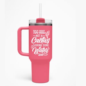 May include: A pink travel mug with a white handle and a white design that says "You are not a cactus, drink some water".