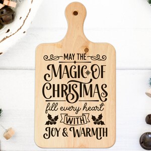 Puede incluir: Una tabla de cortar de madera con un dise&ntilde;o navide&ntilde;o. El texto dice "Que la magia de la Navidad llene cada coraz&oacute;n de alegr&iacute;a y calidez".