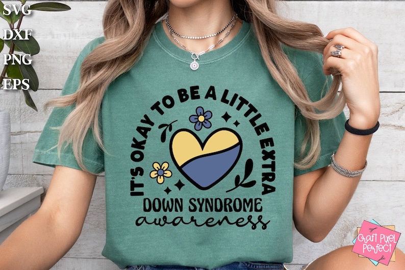 May include: A teal t-shirt with a black circle design that says "It's okay to be a little extra Down Syndrome awareness". The design also includes a yellow and blue heart with flowers.