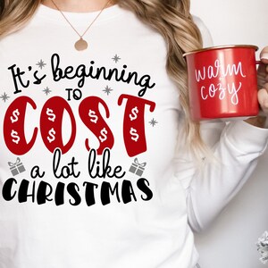 May include: White long-sleeve shirt with the text "It's beginning to cost a lot like Christmas" in black and red lettering. The word "cost" is in red and has dollar signs around it.