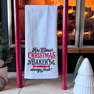 K&ouml;nnte beinhalten: Ein wei&szlig;es Geschirrtuch mit dem Text "Mrs. Claus's Christmas Bakery always fresh!" in schwarzer und roter Schrift. Das Handtuch h&auml;ngt an einer roten Holzleiter vor einem Kamin.