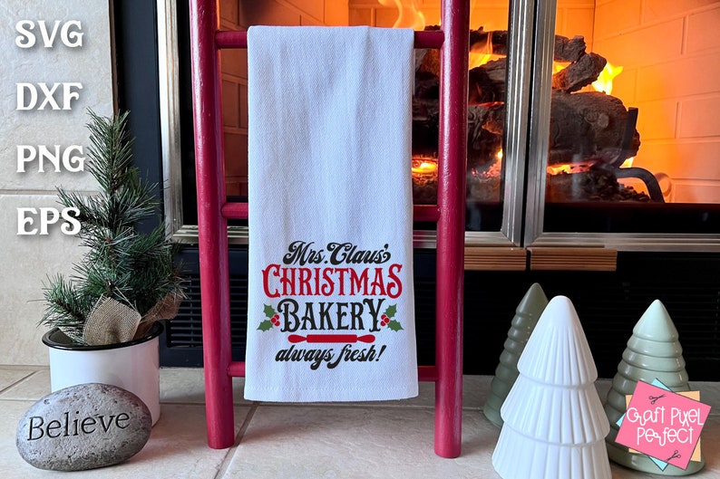 K&ouml;nnte beinhalten: Ein wei&szlig;es Geschirrtuch mit dem Text "Mrs. Claus's Christmas Bakery always fresh!" in schwarzer und roter Schrift. Das Handtuch h&auml;ngt an einer roten Holzleiter vor einem Kamin.
