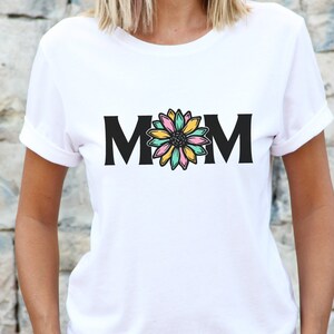 May include: White t-shirt with black text that reads "MOM" and a colorful flower graphic in the center.