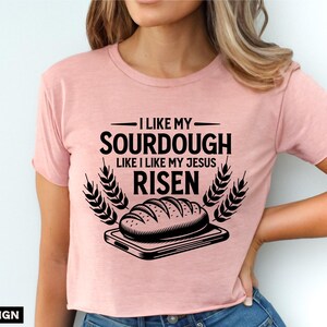May include: A pink t-shirt with a black graphic design that says "I like my sourdough like I like my Jesus risen" with a drawing of a loaf of bread on a cutting board.
