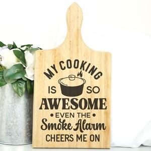 May include: A wooden cutting board with the text "My cooking is so awesome even the smoke alarm cheers me on".