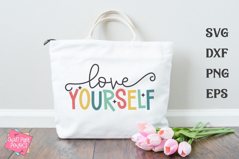 May include: A white tote bag with the text "love yourself" in colorful letters. The letters are in a rainbow color scheme. The bag is sitting on a wooden surface with a bouquet of pink tulips.