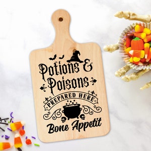 Pu&ograve; includere: Un tagliere di legno con un design in bianco e nero che dice "Pozioni e Veleni Preparati Qui Buon appetito". Il design include un cappello da strega, un calderone e pipistrelli.