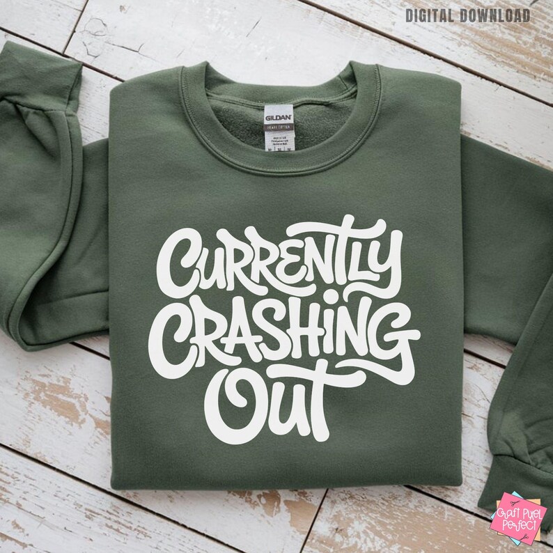 May include: An olive green sweatshirt with the words "Currently Crashing Out" in white, stylized lettering. The sweatshirt is folded, with the sleeves visible. The words "Digital Download" are in the top right corner.