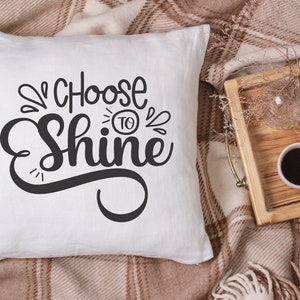 May include: White cushion with black text that reads "Choose to Shine" on a brown and white checked blanket.