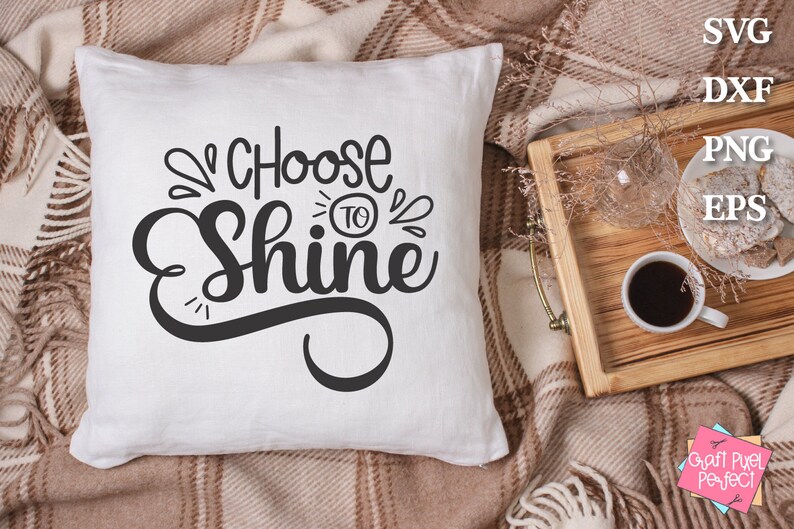 May include: White cushion with black text that reads "Choose to Shine" on a brown and white checked blanket.