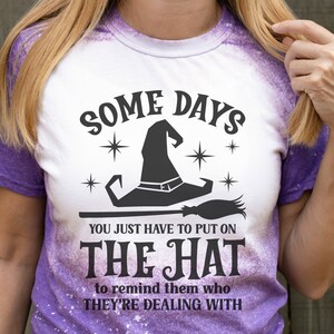 May include: A white t-shirt with a purple tie-dye design featuring a black witch hat and broom with the text "Some Days You Just Have To Put On The Hat To Remind Them Who They're Dealing With".