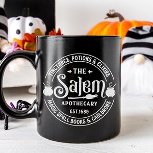 Pu&ograve; includere: Tazza in ceramica nera con un design bianco che recita "Tinctures, Potions & Elixirs The Salem Apothecary Est. 1689 Magic Spell Books & Cauldrons".