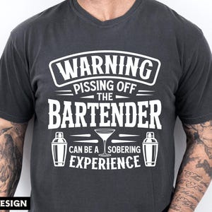 May include: Dark gray t-shirt with a humorous graphic in white. The text reads "WARNING PISSING OFF THE BARTENDER CAN BE A SOBERING EXPERIENCE" with cocktail shaker and martini glass illustrations. Digital design.