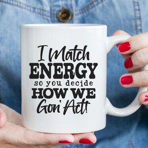 May include: White ceramic mug with the text "I Match ENERGY so you decide HOW WE Gon' Act!" in black. The mug is held by a person with red nail polish. The mug is a standard size for coffee or tea.