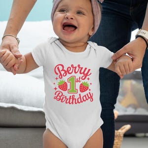 May include: A white onesie with a red and green graphic that says "Berry 1st Birthday". The onesie is being worn by a baby girl.