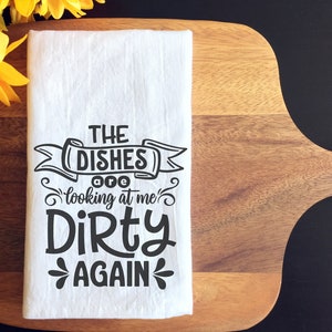 May include: White kitchen towel with the text "THE DISHES are looking at me DIRTY AGAIN" in black. The towel is folded and placed on a wooden cutting board. Yellow sunflowers are in the background.