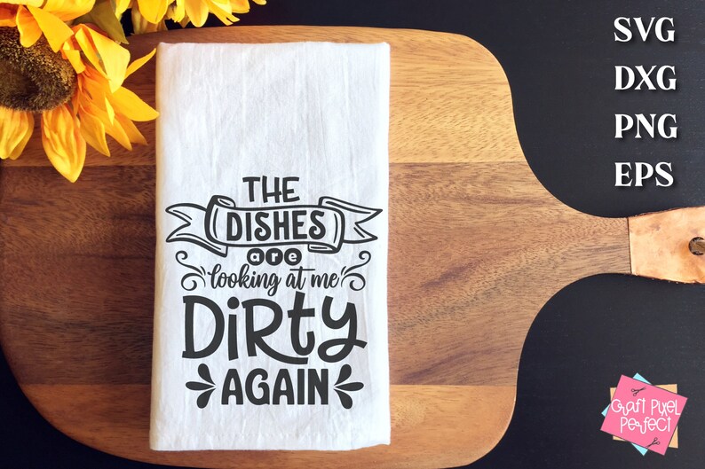 May include: White kitchen towel with the text "THE DISHES are looking at me DIRTY AGAIN" in black. The towel is folded and placed on a wooden cutting board. Yellow sunflowers are in the background.