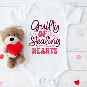 May include: White baby onesie with the text "Guilty of Stealing Hearts" in pink and maroon lettering with pink hearts and an arrow.