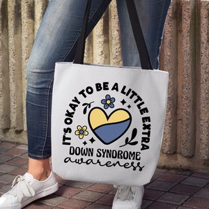 May include: A white tote bag with a black handle and a blue and yellow heart with flowers and the text "It's okay to be a little extra Down Syndrome awareness".