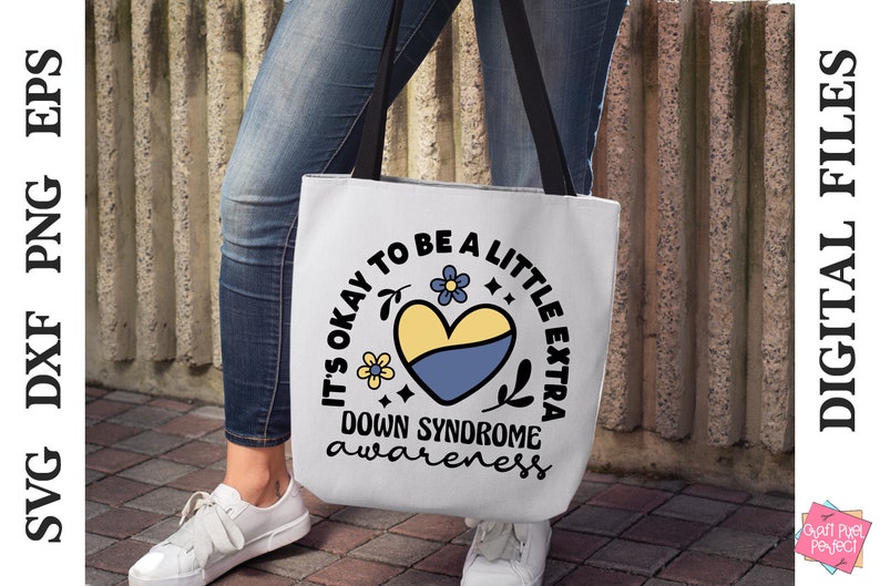 May include: A white tote bag with a black handle and a blue and yellow heart with flowers and the text "It's okay to be a little extra Down Syndrome awareness".