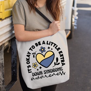 May include: A white tote bag with a black strap and a yellow and blue heart design. The text on the bag reads "It's okay to be a little extra Down Syndrome awareness".