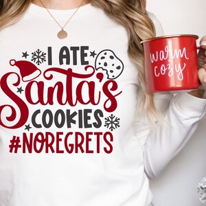 May include: White long-sleeve shirt with red text that says "I ate Santa's cookies #noregrets". The shirt also has a red Santa hat and a black and white cookie graphic. A person is holding a red mug with white text that says "warm cozy".
