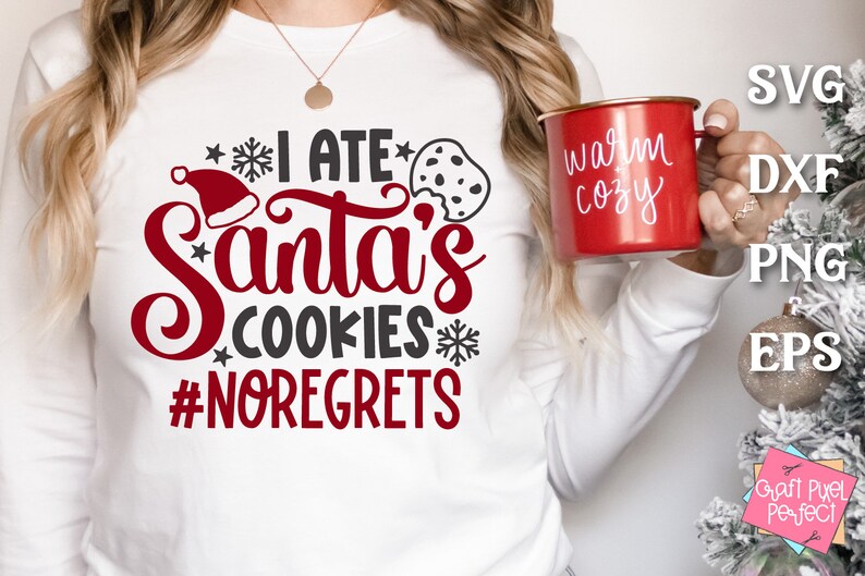 May include: White long-sleeve shirt with red text that says "I ate Santa's cookies #noregrets". The shirt also has a red Santa hat and a black and white cookie graphic. A person is holding a red mug with white text that says "warm cozy".