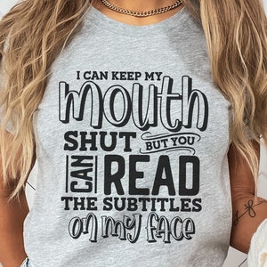 K&ouml;nnte beinhalten: Hellgraues T-Shirt mit dem schwarzen Text "I CAN KEEP MY MOUTH SHUT BUT YOU CAN READ THE SUBTITLES ON MY FACE". Ein l&auml;ssiges Kleidungsst&uuml;ck.