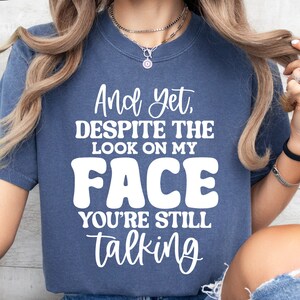 May include: A blue t-shirt with white text that reads "And yet, despite the look on my face you're still talking".