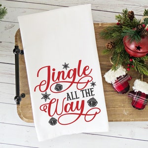 May include: White kitchen towel with the text "Jingle All The Way" in red cursive lettering with black snowflake and bell accents.