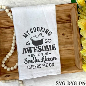 May include: A white tea towel with black text that reads "My cooking is so awesome even the smoke alarm cheers me on". The text is in a retro font style. A black and white illustration of a pot with steam coming out of it is above the word "awesome".