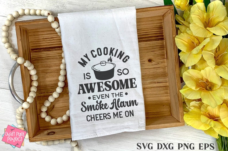 May include: A white tea towel with black text that reads "My cooking is so awesome even the smoke alarm cheers me on". The text is in a retro font style. A black and white illustration of a pot with steam coming out of it is above the word "awesome".