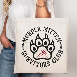Pu&ograve; includere: Una borsa di tela bianca con un design circolare nero che dice "Murder Mitten Survivors Club" con un'impronta di zampa nera con graffi rossi all'interno del cerchio.