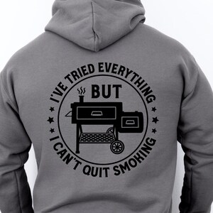 May include: A gray hoodie with a black circle graphic on the back. The graphic features a smoker with the text "I've tried everything but I can't quit smoking".